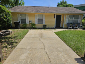 aquaTech pressure washing cleaning a driveway and sidewalk near a yellow house with green grass and seating area