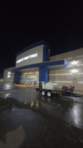 A pressure washing trailer is parked outside a well-lit storefront at night, with the freshly cleaned entrance concrete still wet and reflective