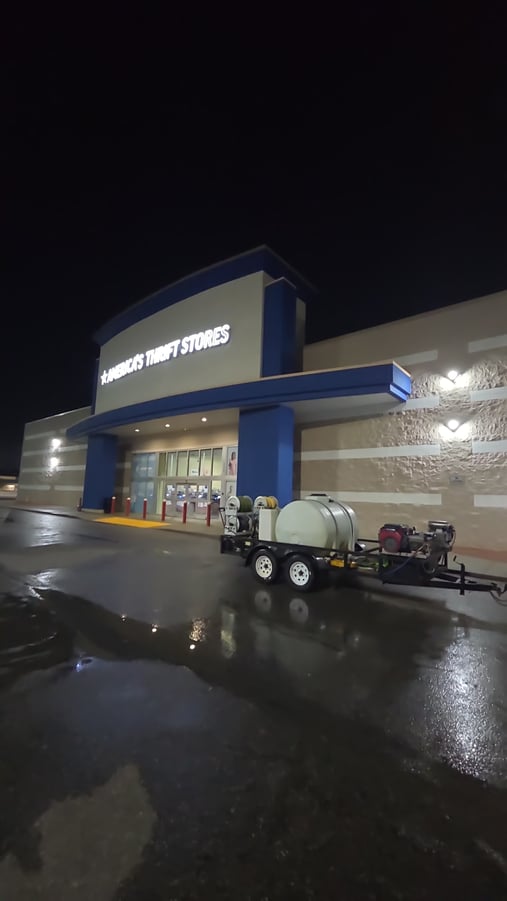 A pressure washing trailer is parked outside a well-lit storefront at night, with the freshly cleaned entrance concrete still wet and reflective
